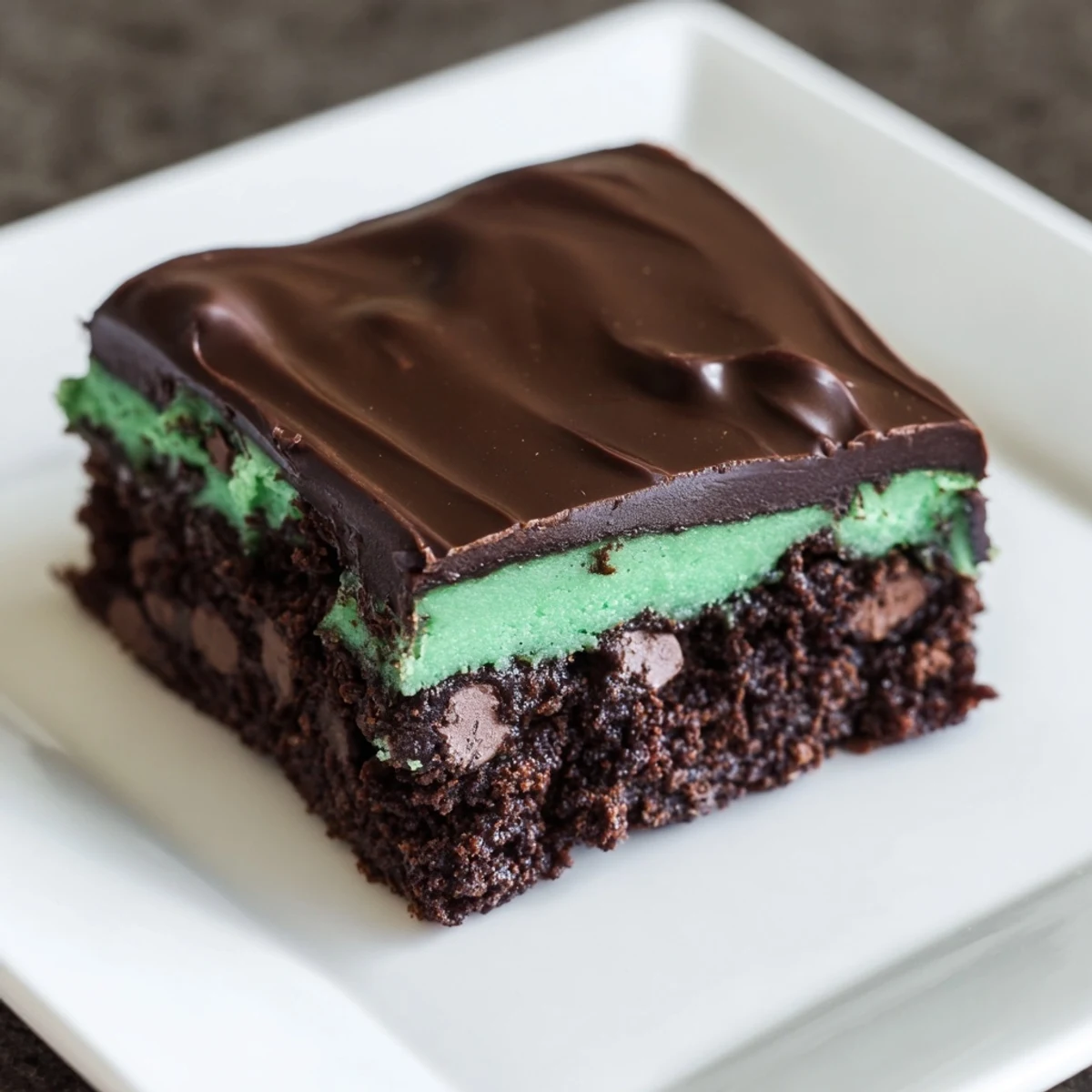 A close-up of Mint Chocolate Chip Brownies showing a fudgy interior, chocolate chips, and a smooth mint green layer topped with glossy chocolate.